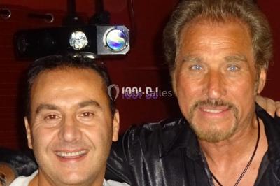 Un homme souriant se tient près d'une table de mixage éclairée, avec un écran projetant une vidéo en arrière-plan.