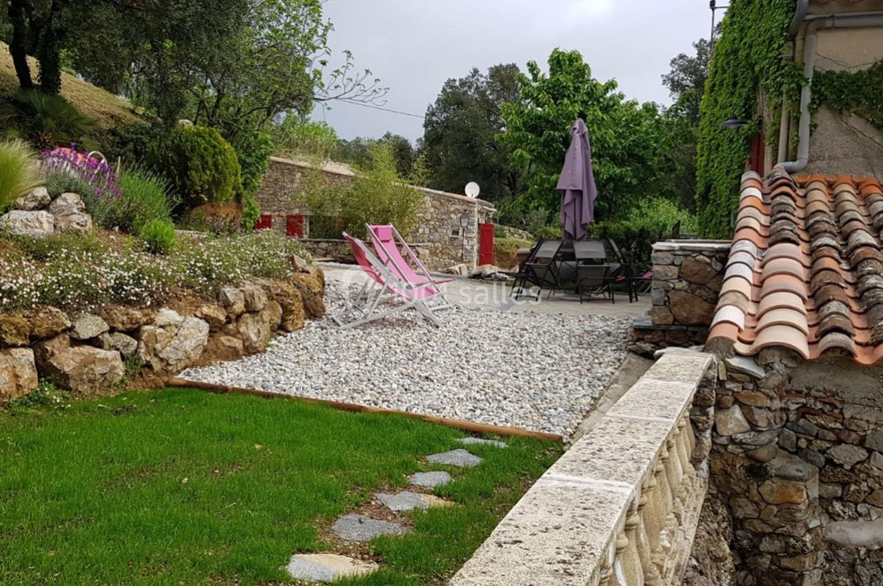 Terrasse extérieure avec chaises longues, parasol et végétation environnante, bordée de murs en pierre et pelouse.