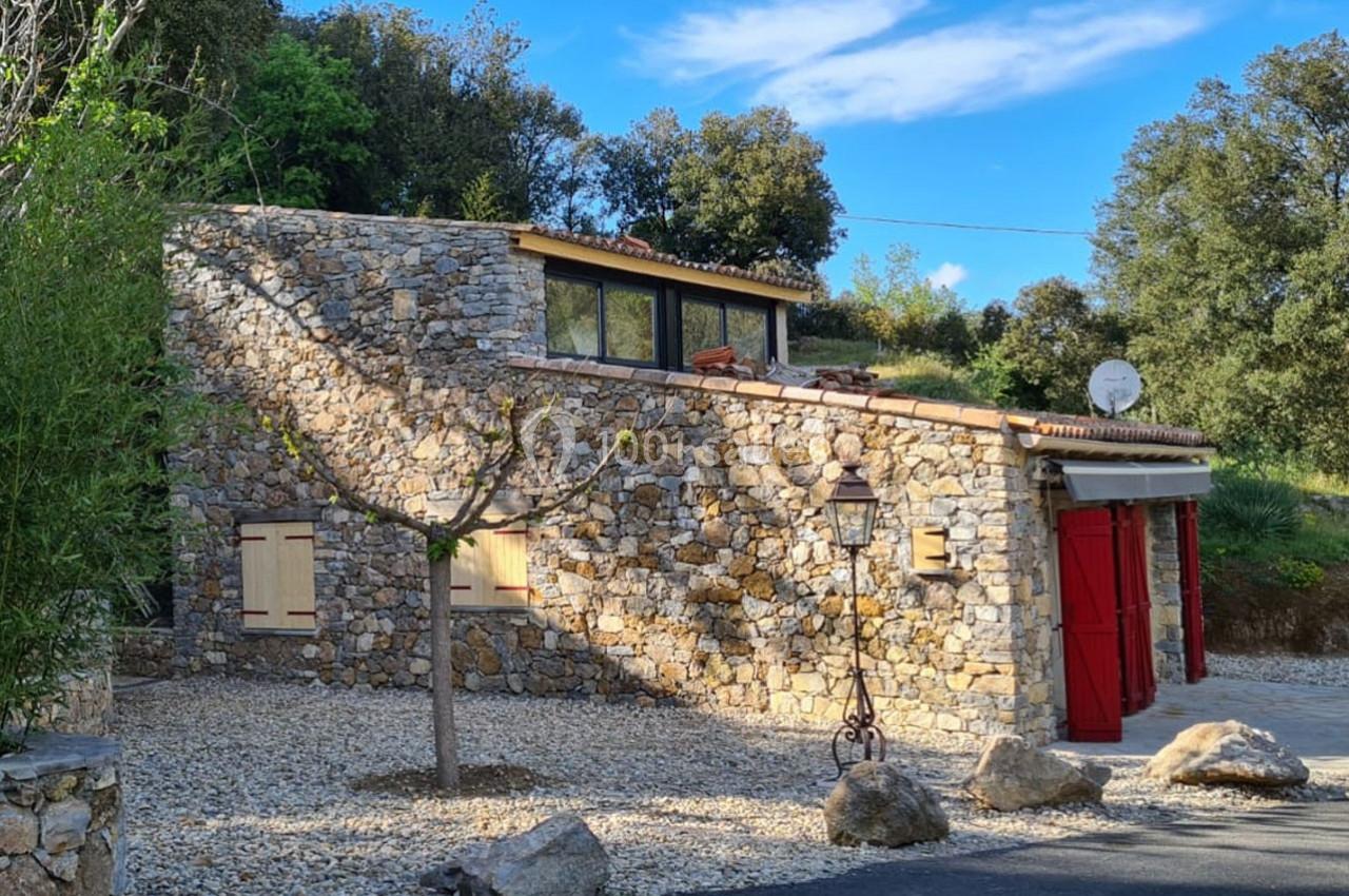 Maison en pierre avec volets rouges, entourée de végétation, sous un ciel bleu clair.