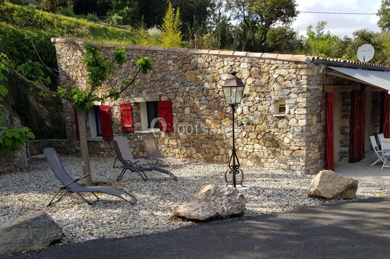 Maison en pierre avec volets rouges, terrasse en gravier, chaises longues et lampadaire, entourée de verdure.