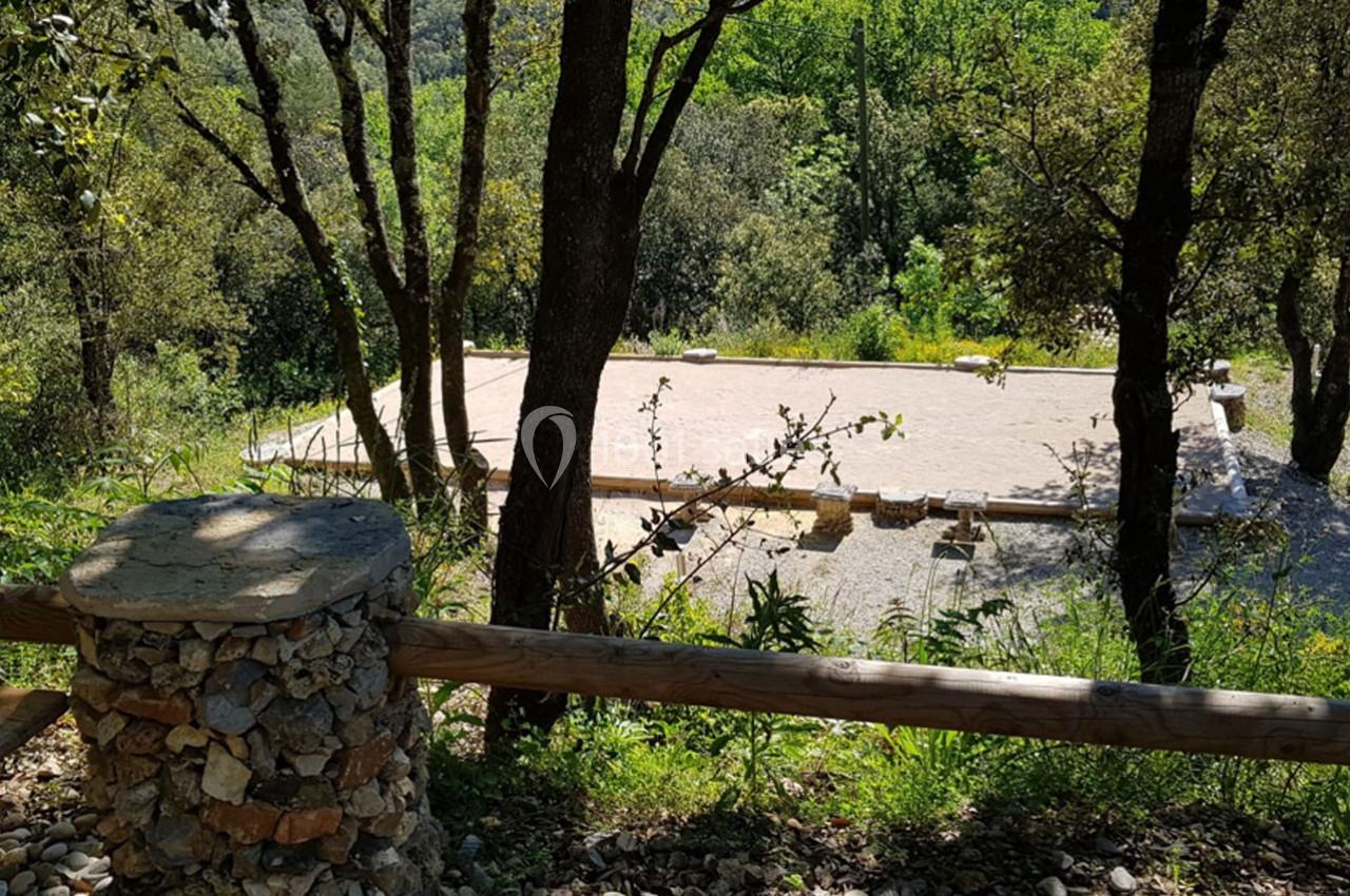 Terrain de pétanque ensoleillé entouré d'arbres et de végétation, avec un muret en pierres au premier plan.
