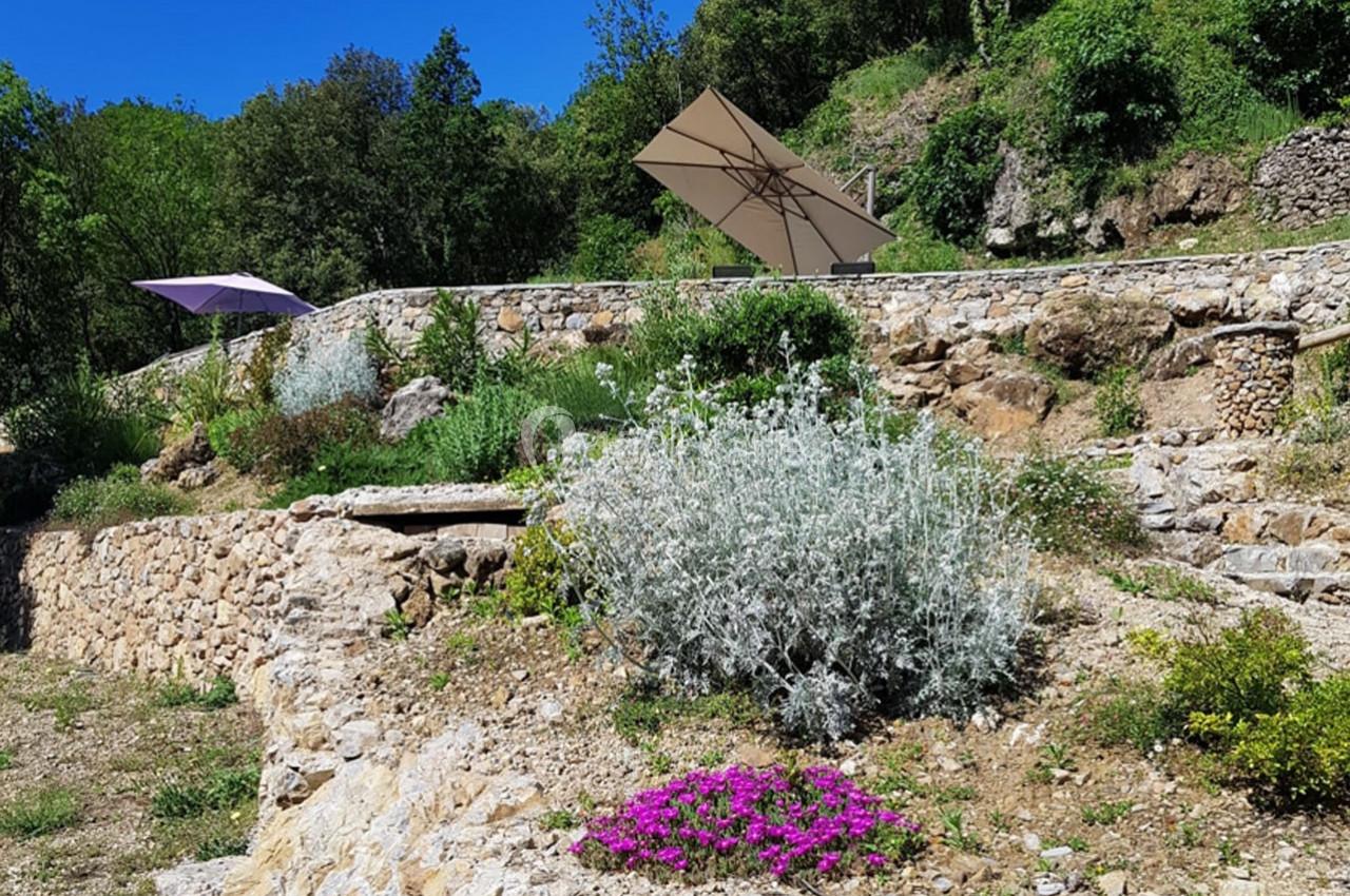 Terrasse en pierre avec végétation méditerranéenne, fleurs violettes et parasols beige et violet sous un ciel bleu.