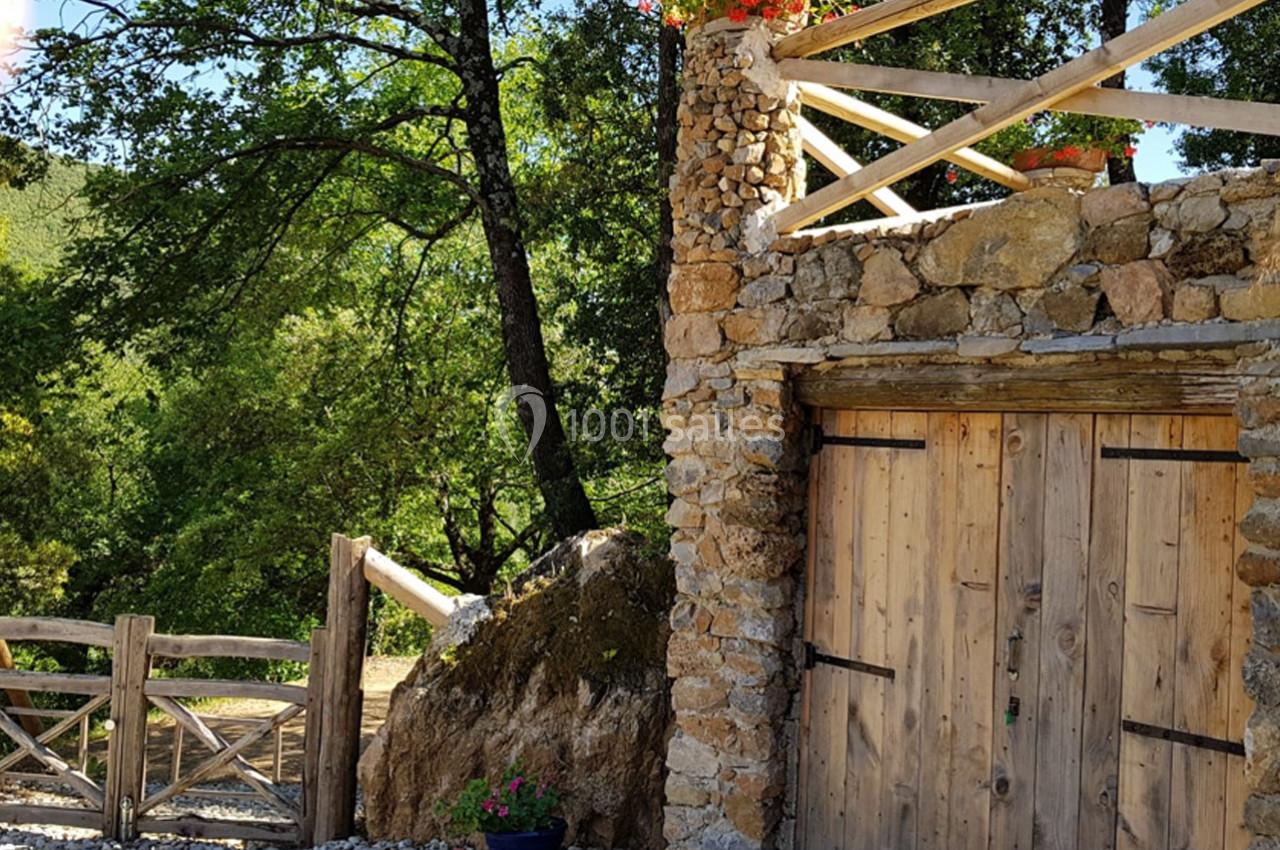 Façade en pierre avec une porte en bois, terrasse fleurie au-dessus, entourée de végétation et d'une clôture en bois.