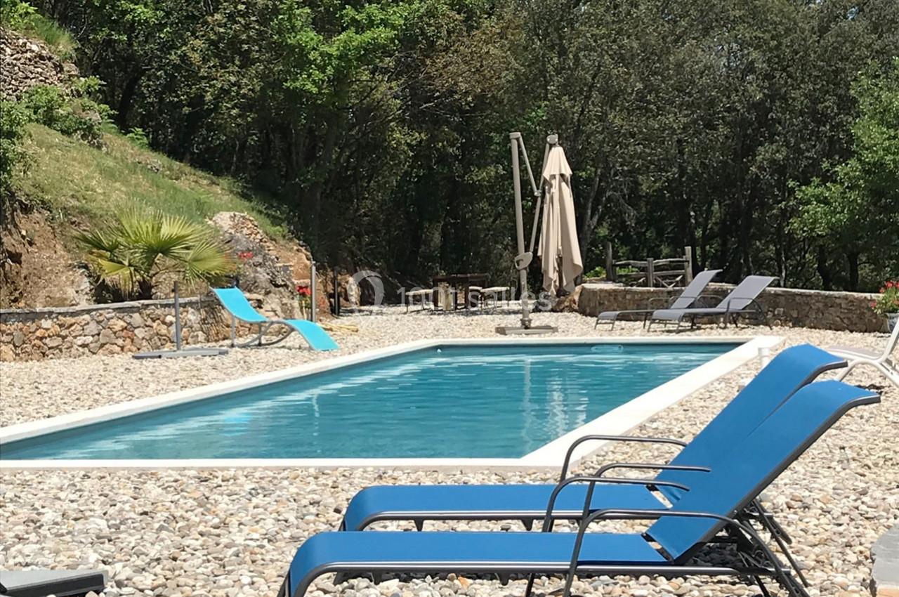 Piscine extérieure entourée de galets, avec transats bleus, parasol beige et végétation en arrière-plan.