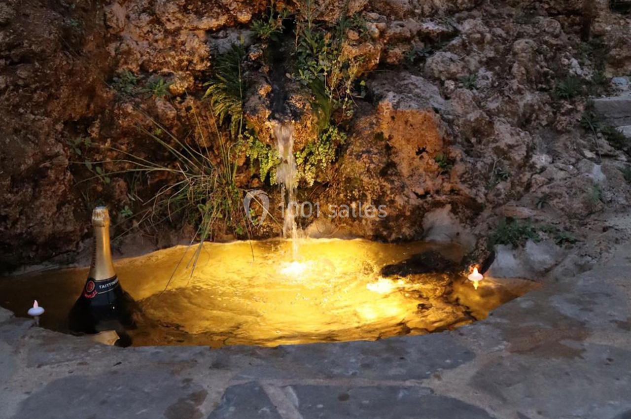 Source d'eau illuminée entourée de pierres, avec une bouteille de champagne et deux bougies posées à proximité.