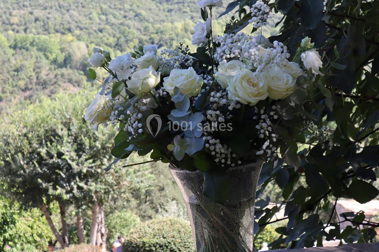 Bouquet de fleurs blanches et vertes dans un vase en verre, posé devant un paysage boisé en arrière-plan.
