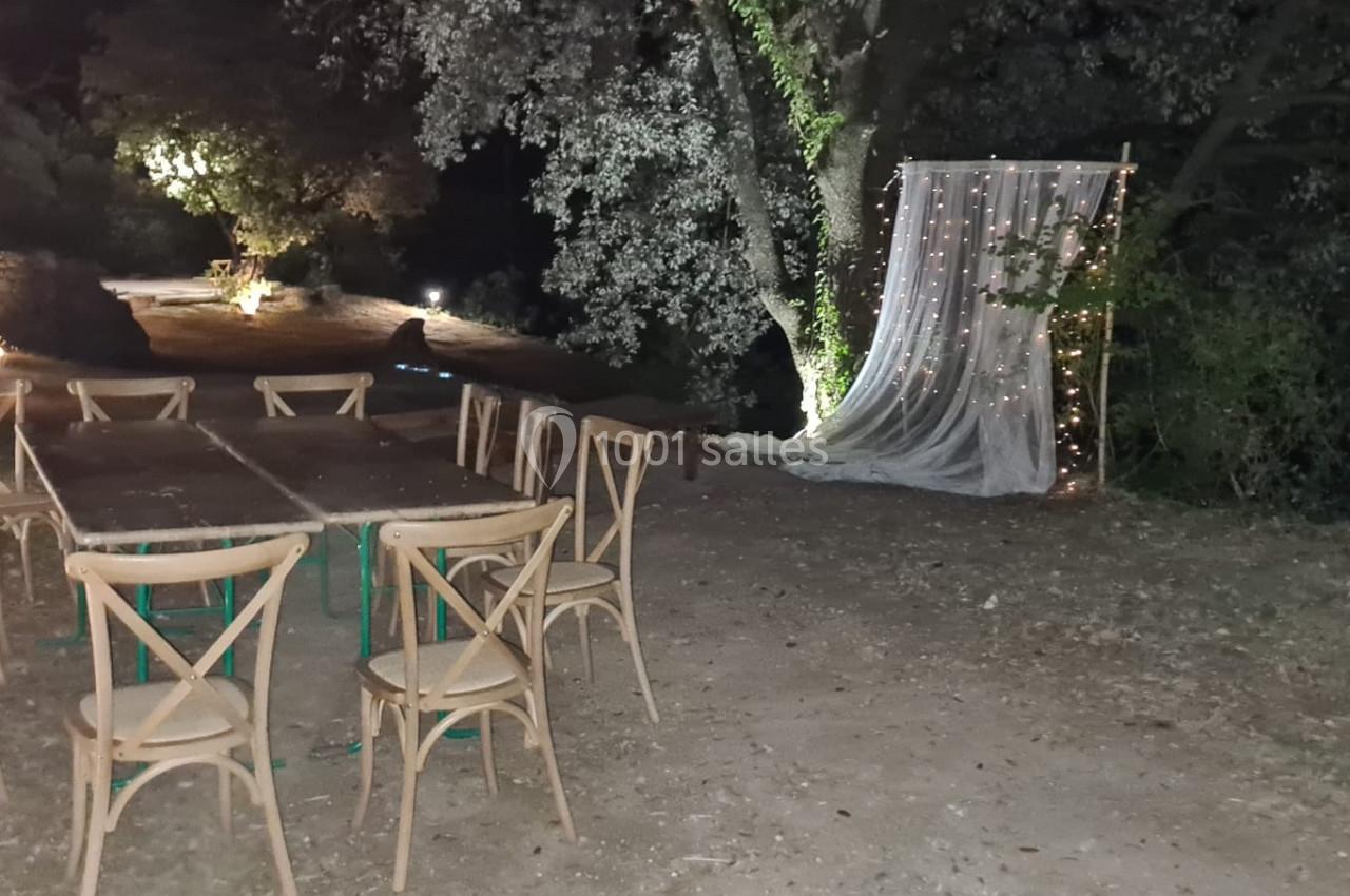 Chaises en bois disposées autour de tables en extérieur, avec un décor lumineux et un rideau blanc sous des arbres la nuit.