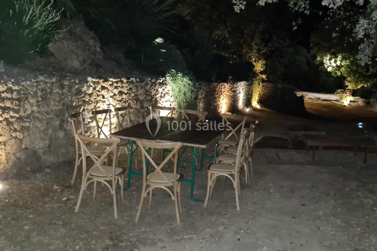 Table et chaises en bois disposées en extérieur, éclairées par des lumières au sol près d'un mur en pierre la nuit.