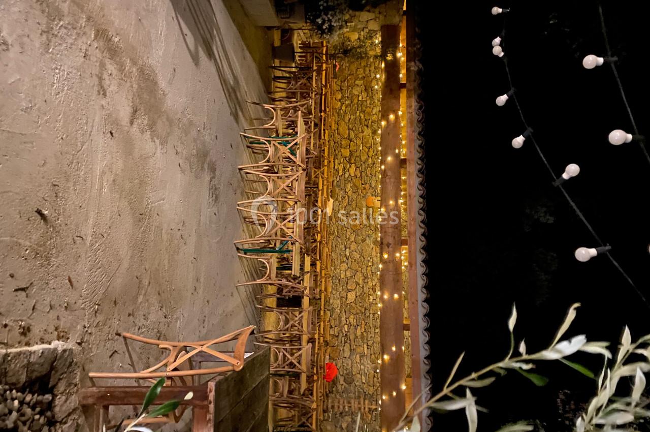 Terrasse extérieure éclairée par des guirlandes lumineuses, avec des chaises en bois alignées contre un mur en pierre.