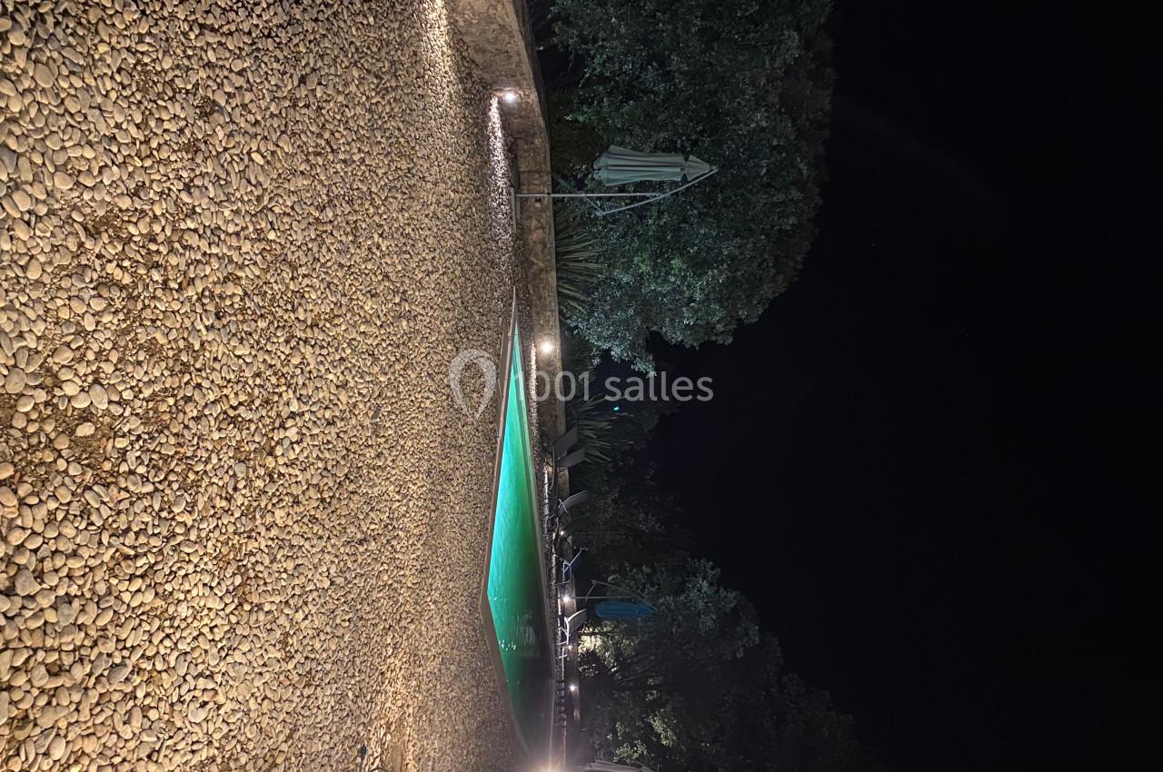 Piscine éclairée entourée de galets et d'arbres dans un jardin, photographiée de nuit.