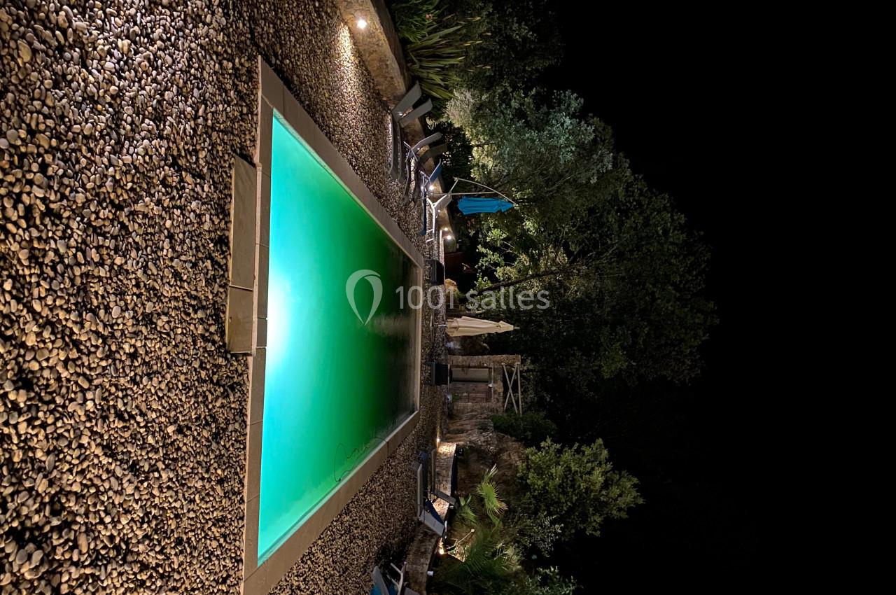 Piscine éclairée de nuit entourée de galets, chaises longues et végétation dense en arrière-plan.