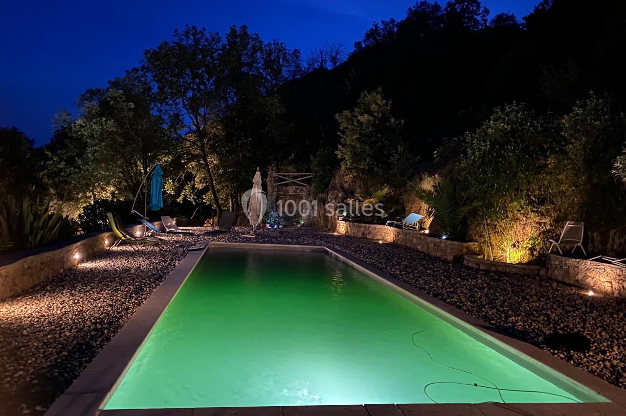 Piscine éclairée entourée de galets et de végétation, avec des chaises longues, sous un ciel nocturne.