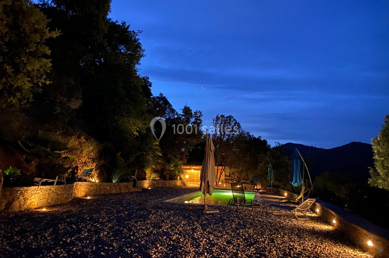 Terrasse éclairée en soirée avec une piscine, des transats et des parasols, entourée de végétation et de collines.