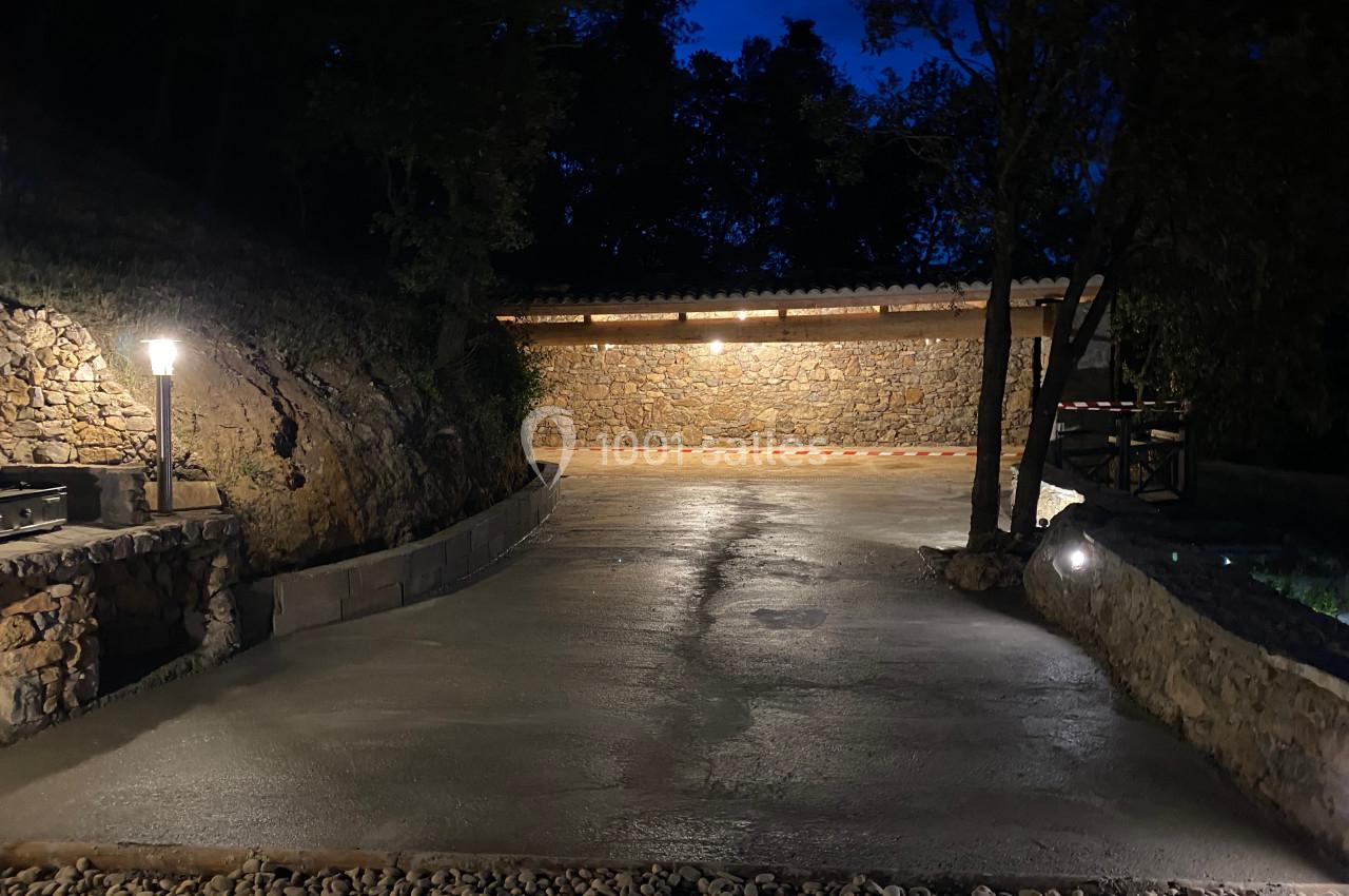 Allée en béton éclairée par des lampadaires, entourée de murs en pierre et d'arbres, de nuit.