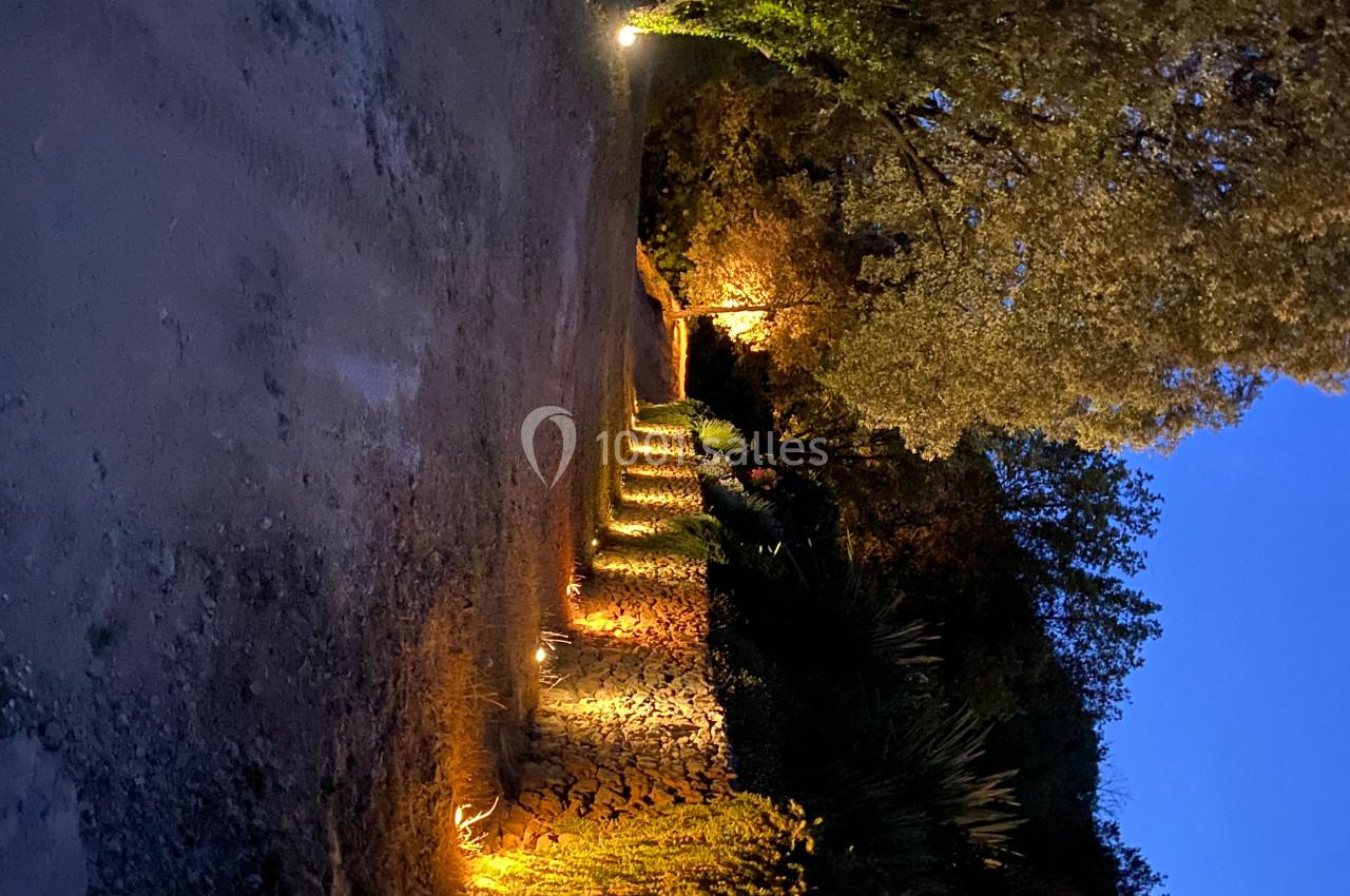 Allée bordée de murs en pierre et de végétation éclairée par des lumières au crépuscule.