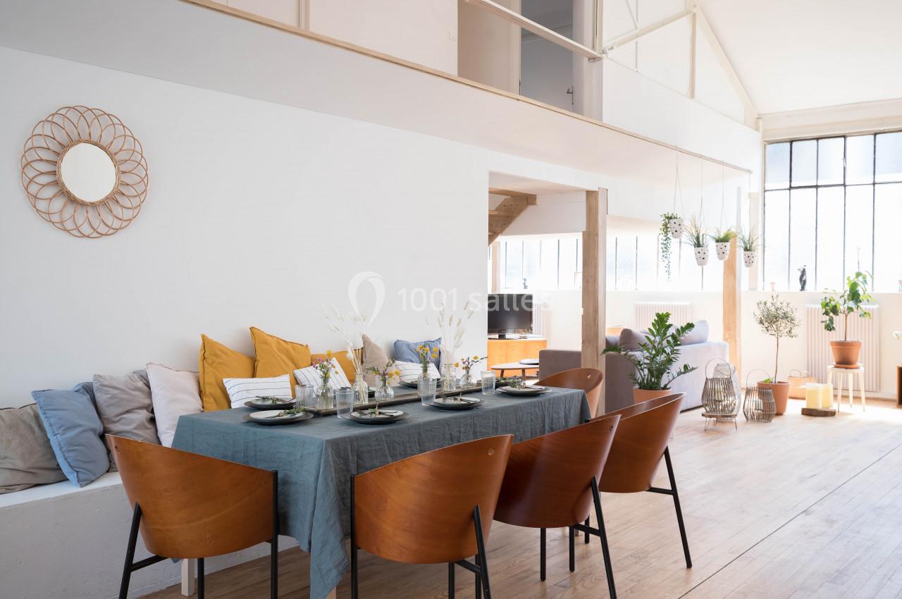 Salle à manger lumineuse avec table dressée, chaises en bois, coussins colorés et plantes près de grandes fenêtres.