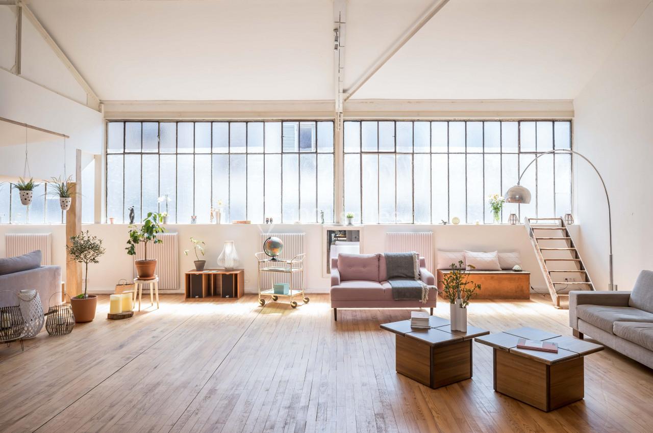 Loft lumineux avec de grandes fenêtres, mobilier moderne, plantes et décoration minimaliste sur parquet en bois clair.