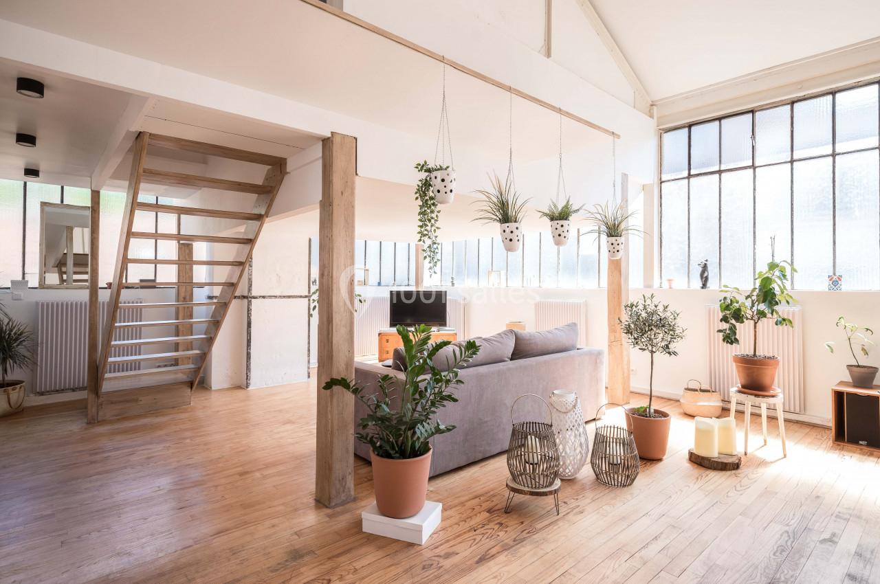 Loft lumineux avec parquet en bois, plantes en pot, canapé gris et grande baie vitrée laissant entrer la lumière naturelle.