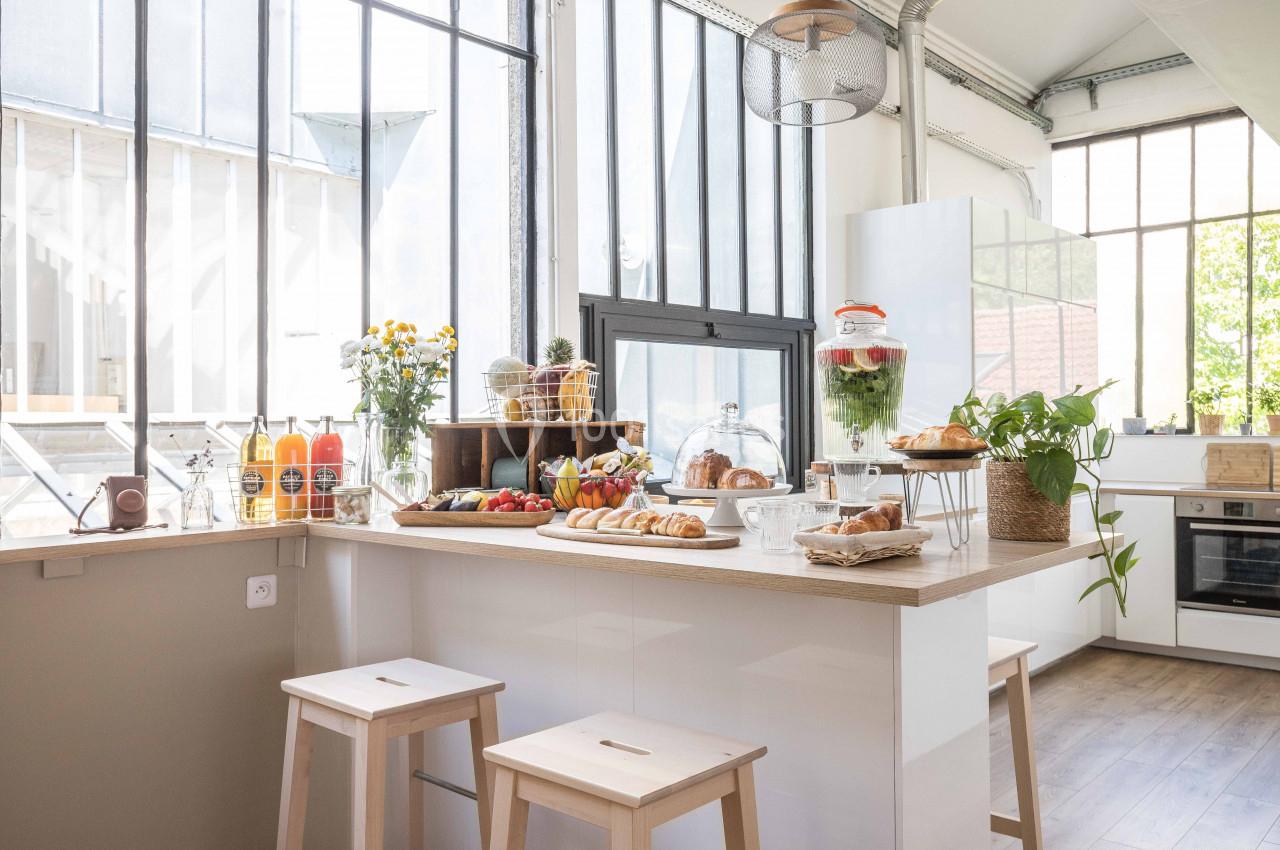 Cuisine lumineuse avec grande verrière, comptoir en bois garni de viennoiseries, fruits, jus et plantes décoratives.