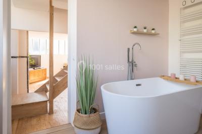 Salle de bain lumineuse avec baignoire blanche, mur rose, plante en pot et vue sur un escalier en bois.