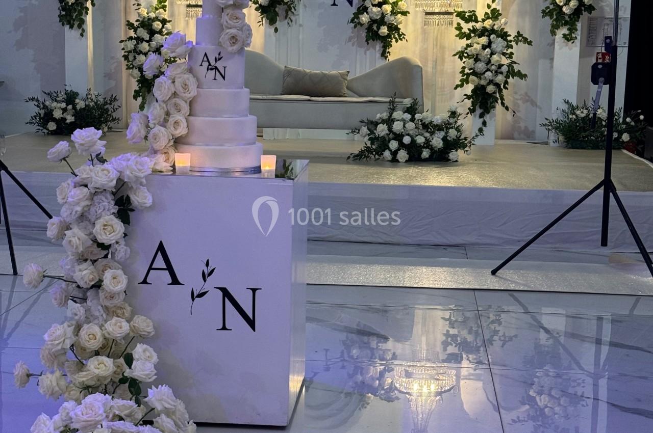 Décor de mariage élégant avec un gâteau blanc, des roses blanches et une scène ornée de fleurs et d'arches lumineuses.