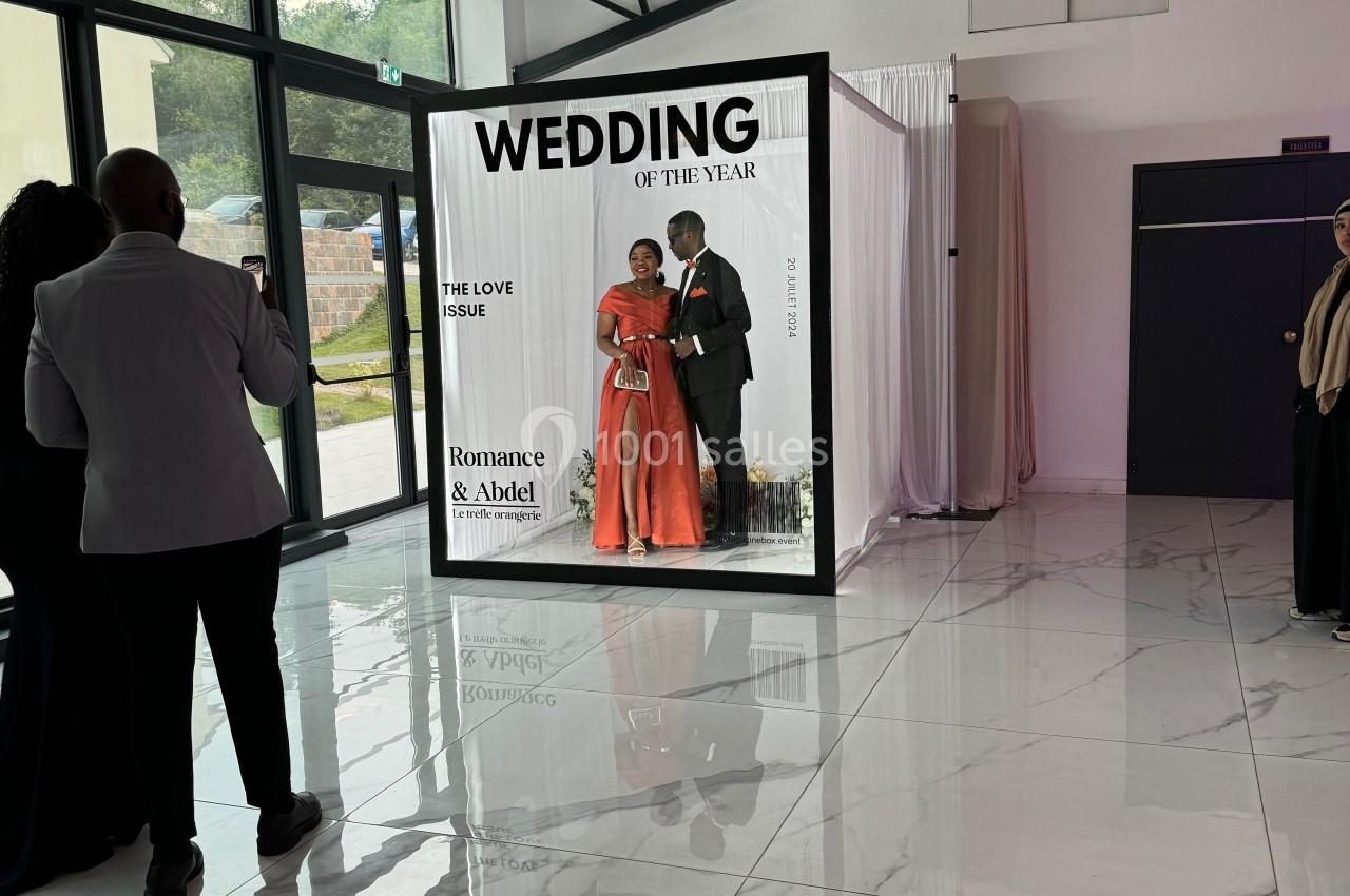 Un couple pose dans une cabine photo décorée pour un mariage, entouré de quelques invités dans une salle lumineuse.