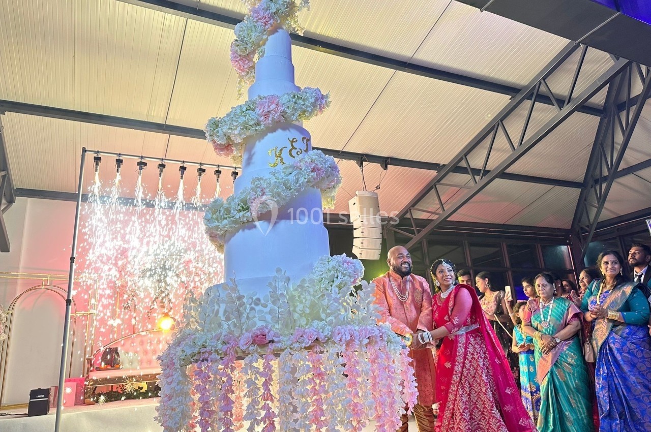 Un couple en tenue traditionnelle coupe un gâteau de mariage blanc décoré de fleurs, entouré d'invités.