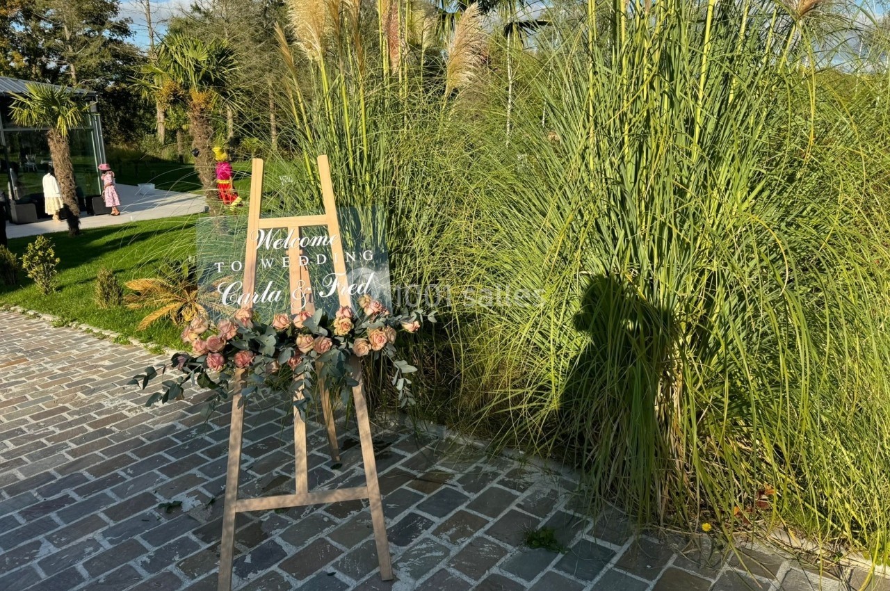 Panneau de bienvenue sur un chevalet décoré de fleurs, placé sur une allée pavée près de hautes herbes.