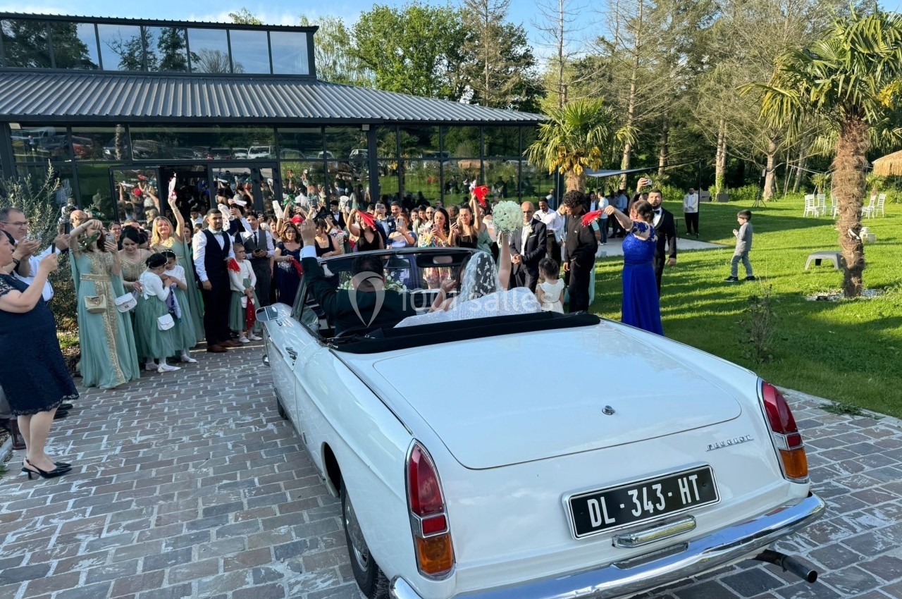 Un couple en tenue de mariage quitte une réception dans une voiture décapotable blanche, entouré d'invités.
