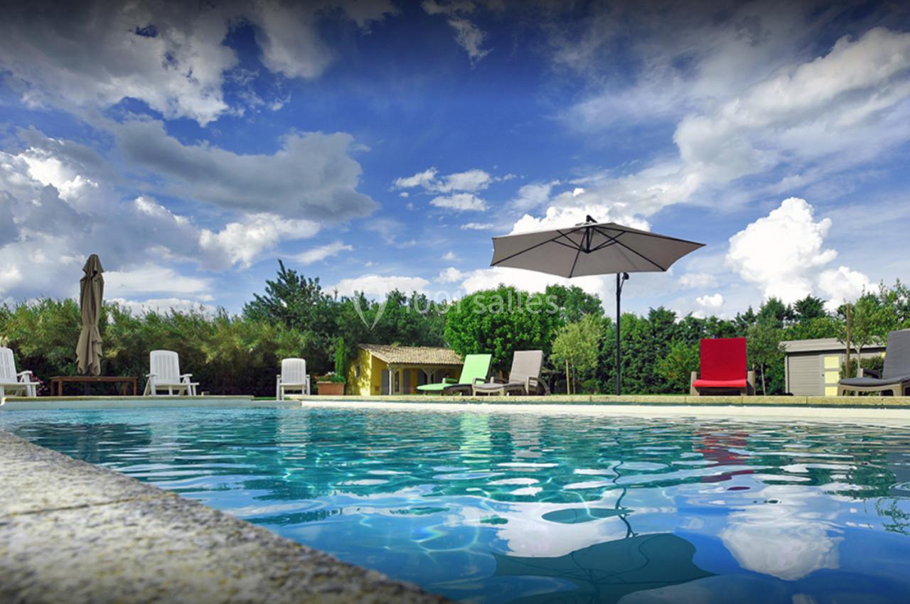 Piscine extérieure entourée de transats colorés, parasol et végétation sous un ciel partiellement nuageux.