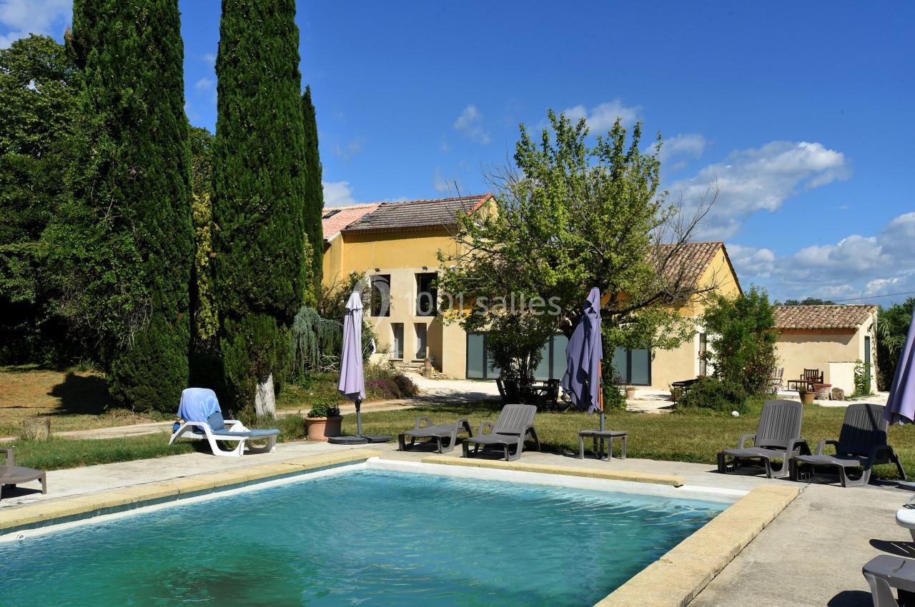 Maison provençale entourée de verdure avec une piscine, des transats et des parasols sous un ciel bleu.