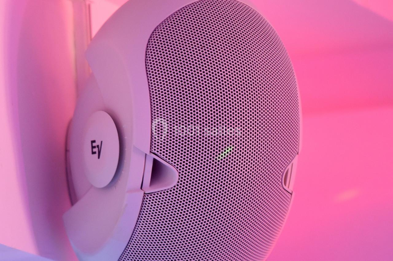 Enceinte murale blanche fixée sous une étagère, éclairée par une lumière rose.