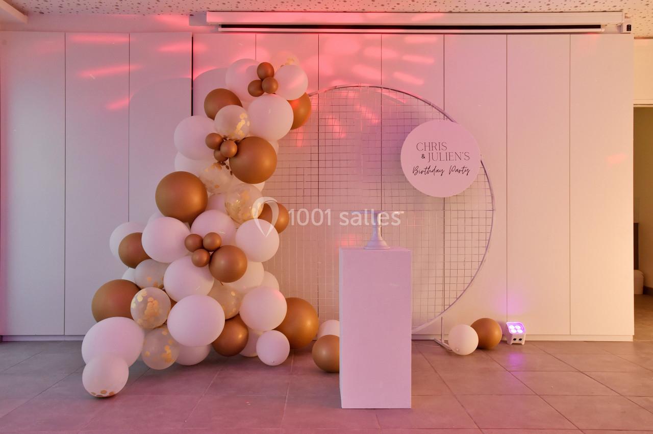 Mur décoratif avec des ballons blancs et dorés, grille métallique ronde et socle blanc dans une salle éclairée.