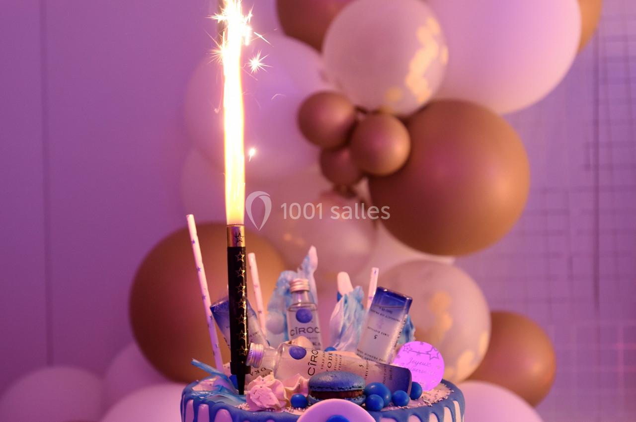 Gâteau décoré avec des bouteilles miniatures, une fontaine lumineuse et des ballons blancs et dorés en arrière-plan.