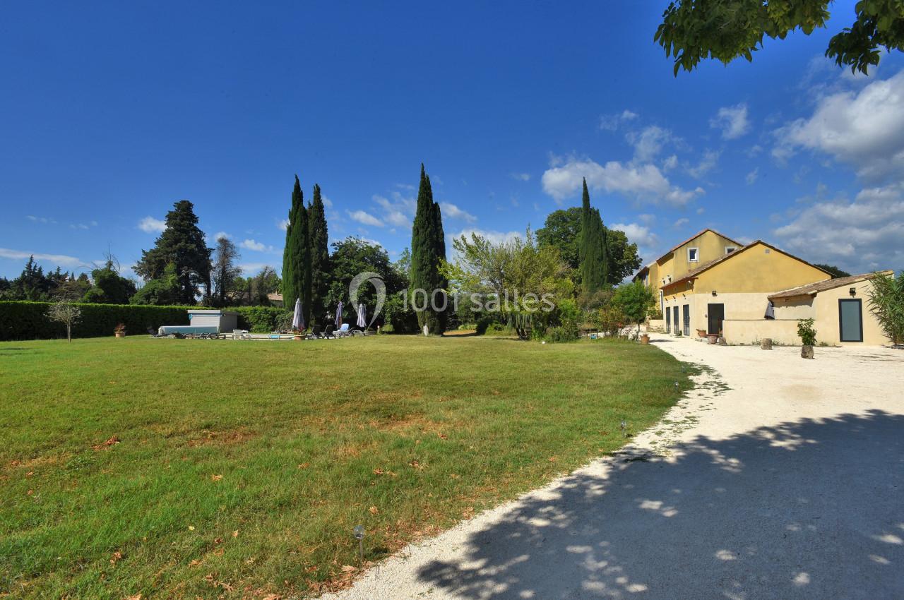 Grande maison jaune avec jardin verdoyant, allée en gravier, cyprès et ciel bleu dégagé en arrière-plan.