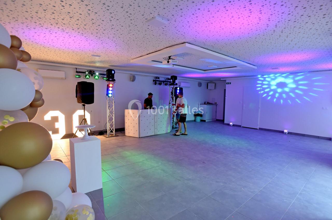 Salle décorée avec des ballons, éclairages colorés et une table de DJ, prête pour un événement festif.