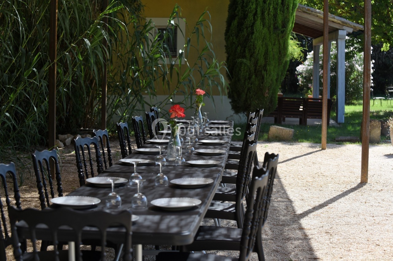 Table extérieure dressée avec assiettes et verres, située dans un jardin ombragé près d'une maison jaune.