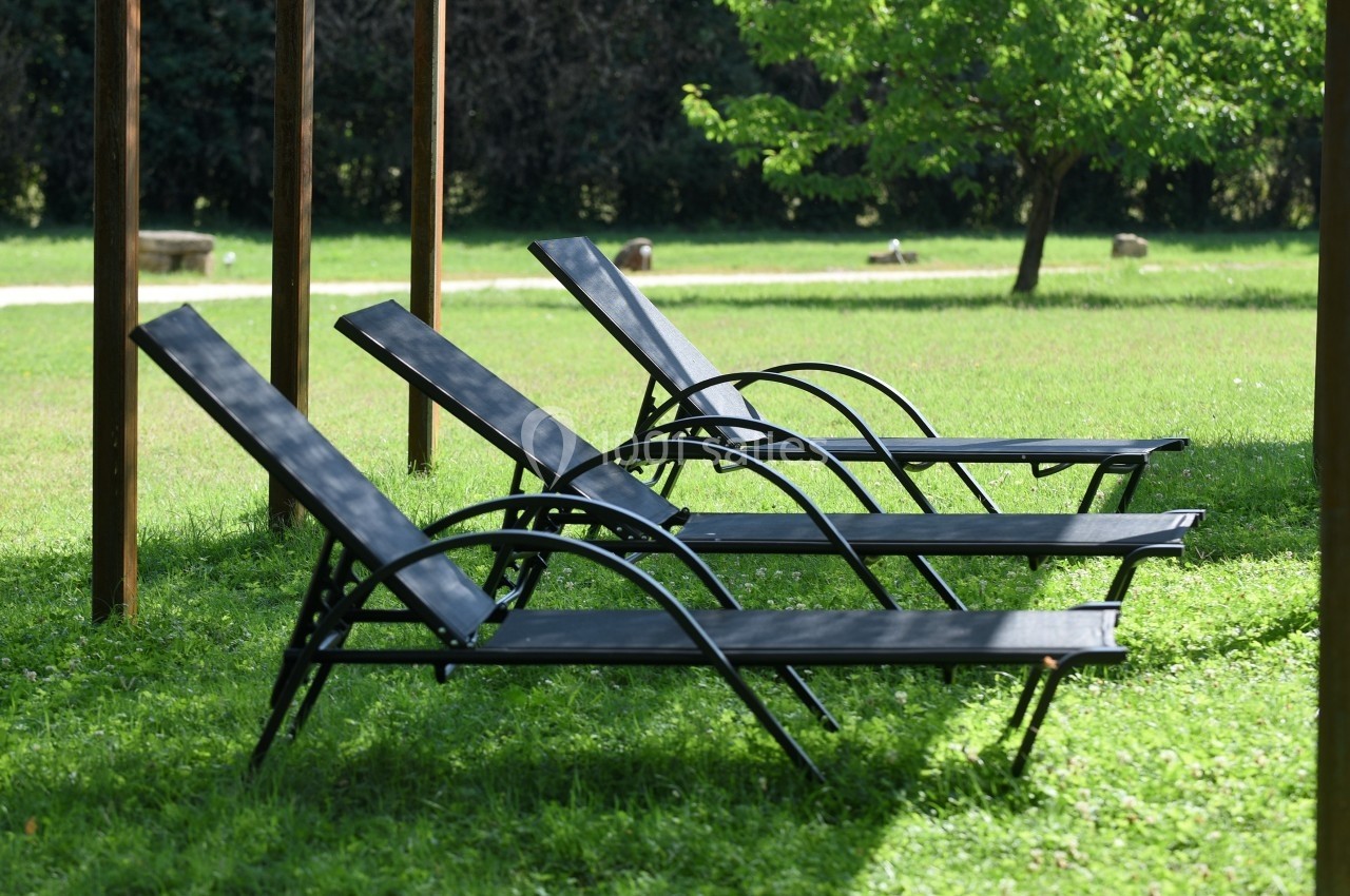 Trois chaises longues noires alignées sur une pelouse verte sous une structure en bois, avec des arbres en arrière-plan.