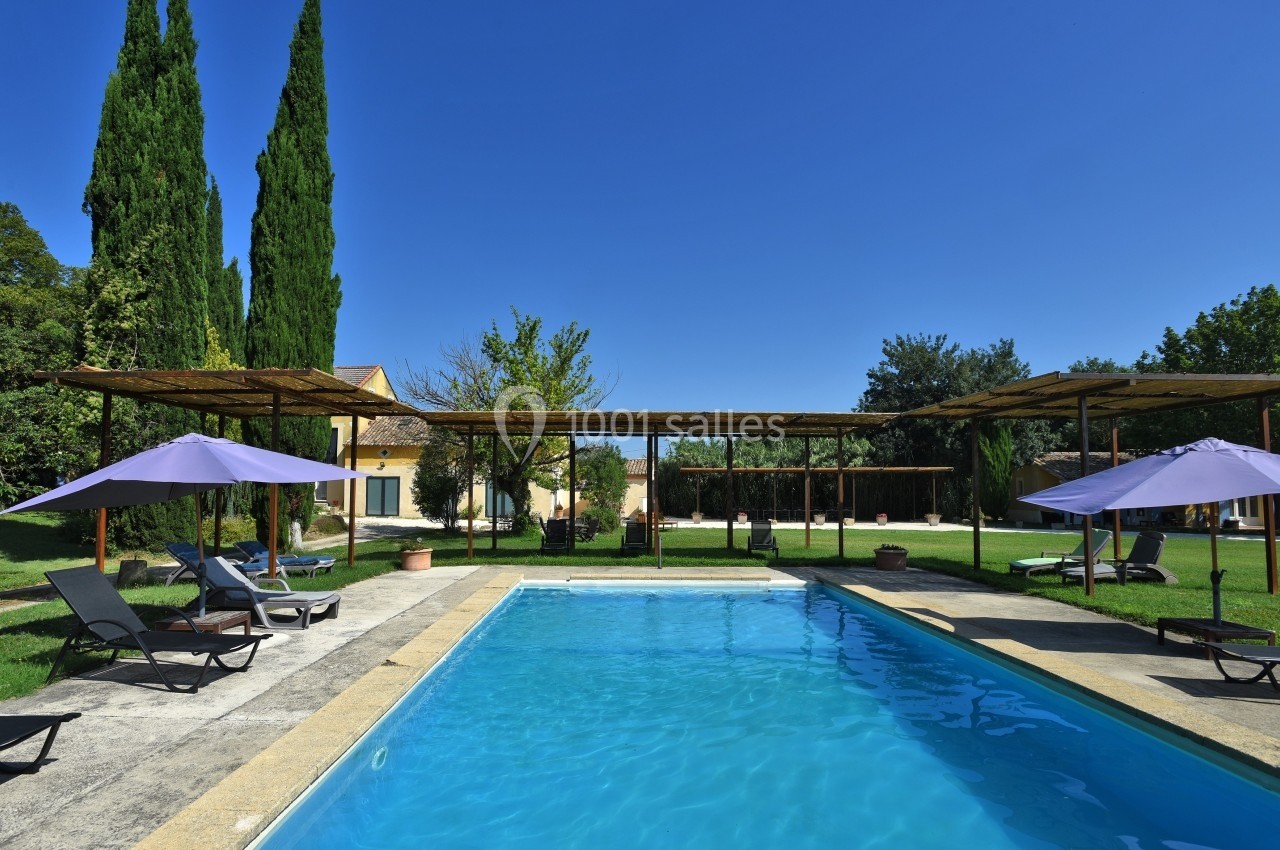 Piscine extérieure entourée de chaises longues, parasols violets et arbres, sous un ciel bleu clair.