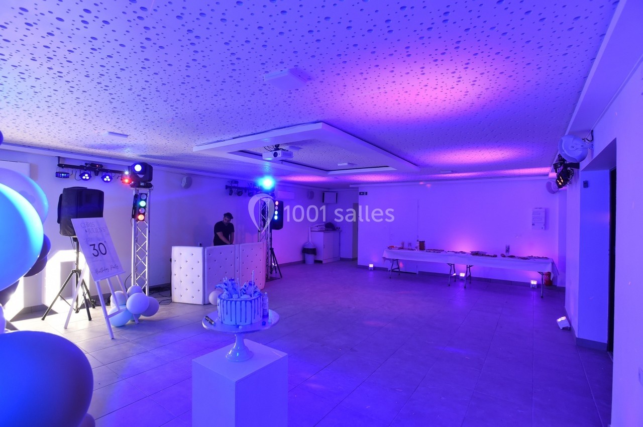 Salle décorée avec éclairage violet, table de buffet au fond, DJ à sa console et gâteau sur un présentoir au premier plan.