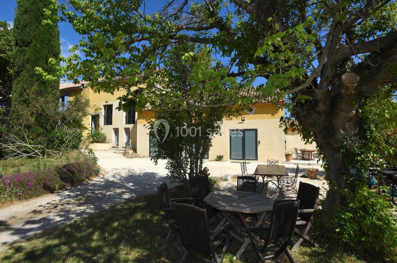 Maison en pierre jaune avec terrasse ombragée par un grand arbre, entourée de mobilier de jardin et de végétation.