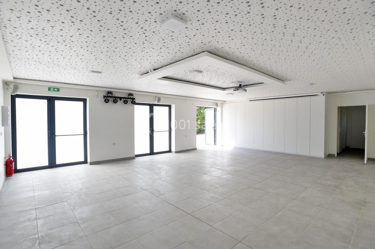 Salle vide avec carrelage gris, plafond acoustique perforé, grandes baies vitrées et porte ouverte sur l'extérieur.