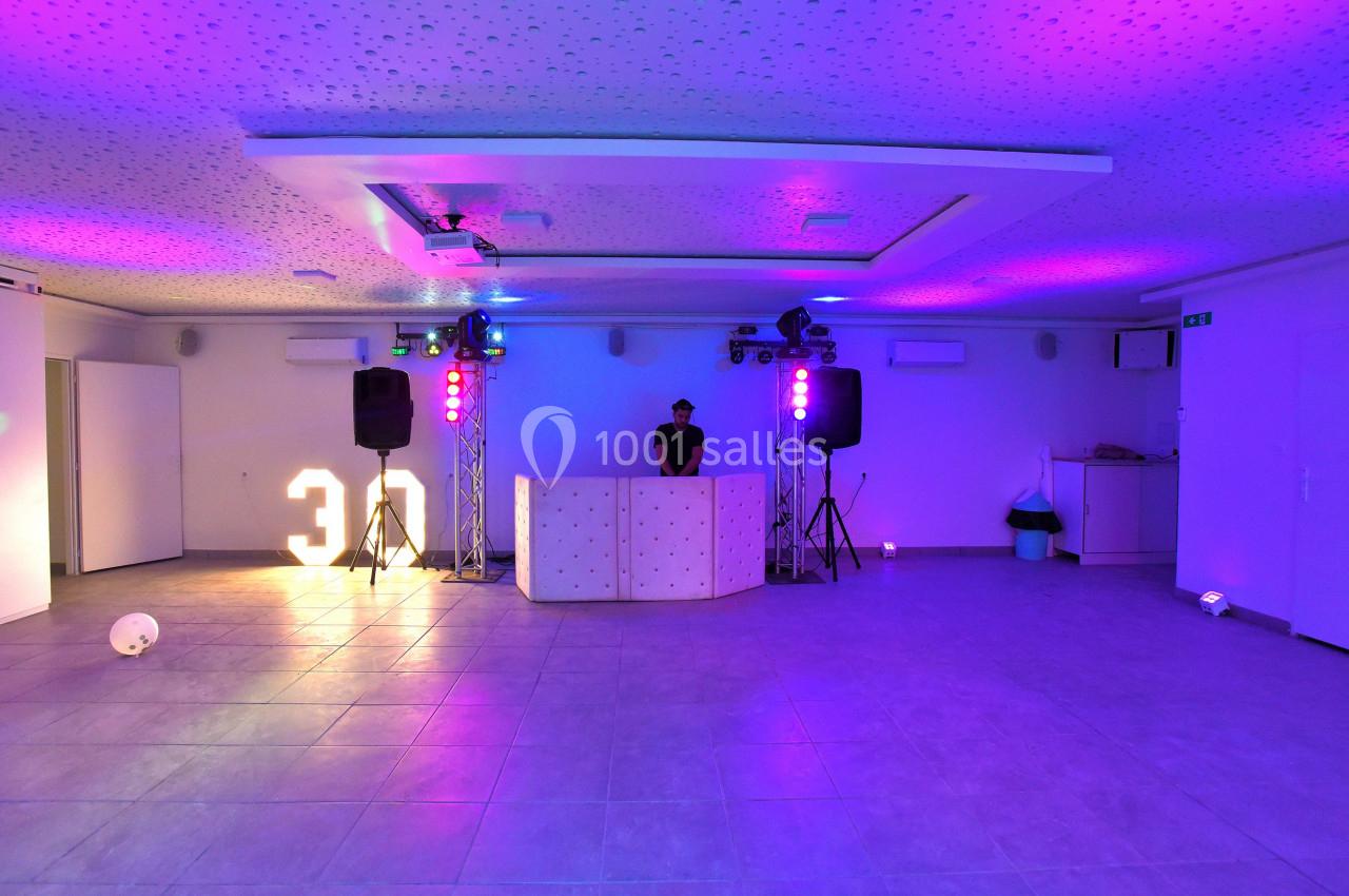 Salle éclairée par des lumières colorées avec un DJ derrière une table, décorée pour un événement festif.