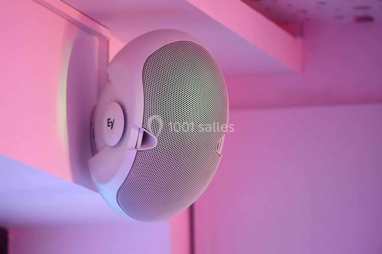 Enceinte murale blanche fixée sous une étagère, éclairée par une lumière rose.