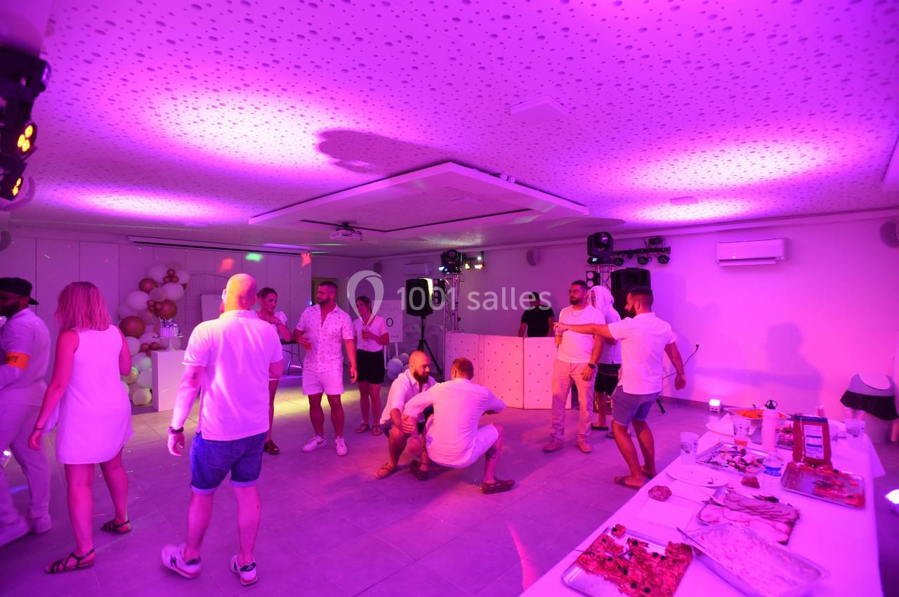 Des personnes dansent et discutent dans une salle éclairée par des lumières violettes, avec des tables et décorations en…