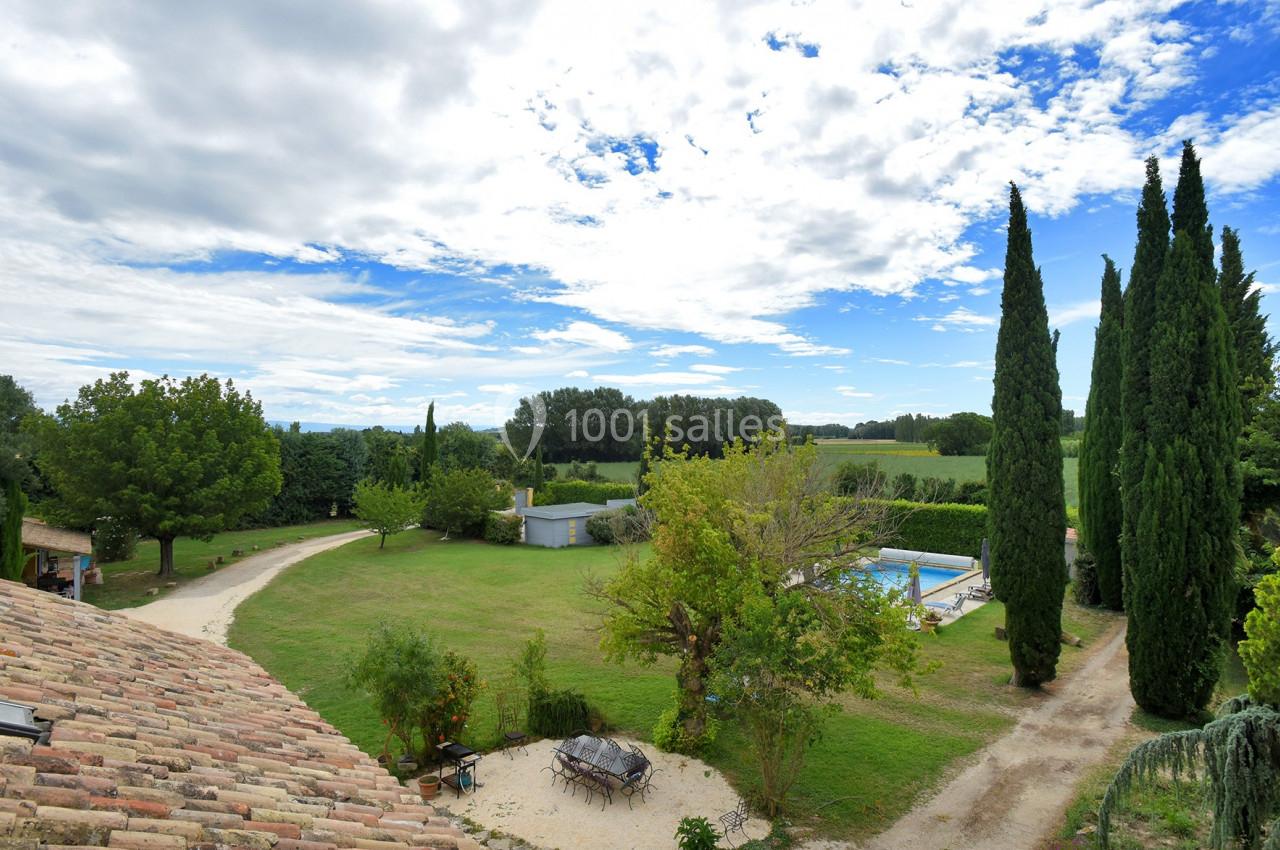 Vue d'un grand jardin avec piscine, allées bordées d'arbres et paysage rural sous un ciel partiellement nuageux.