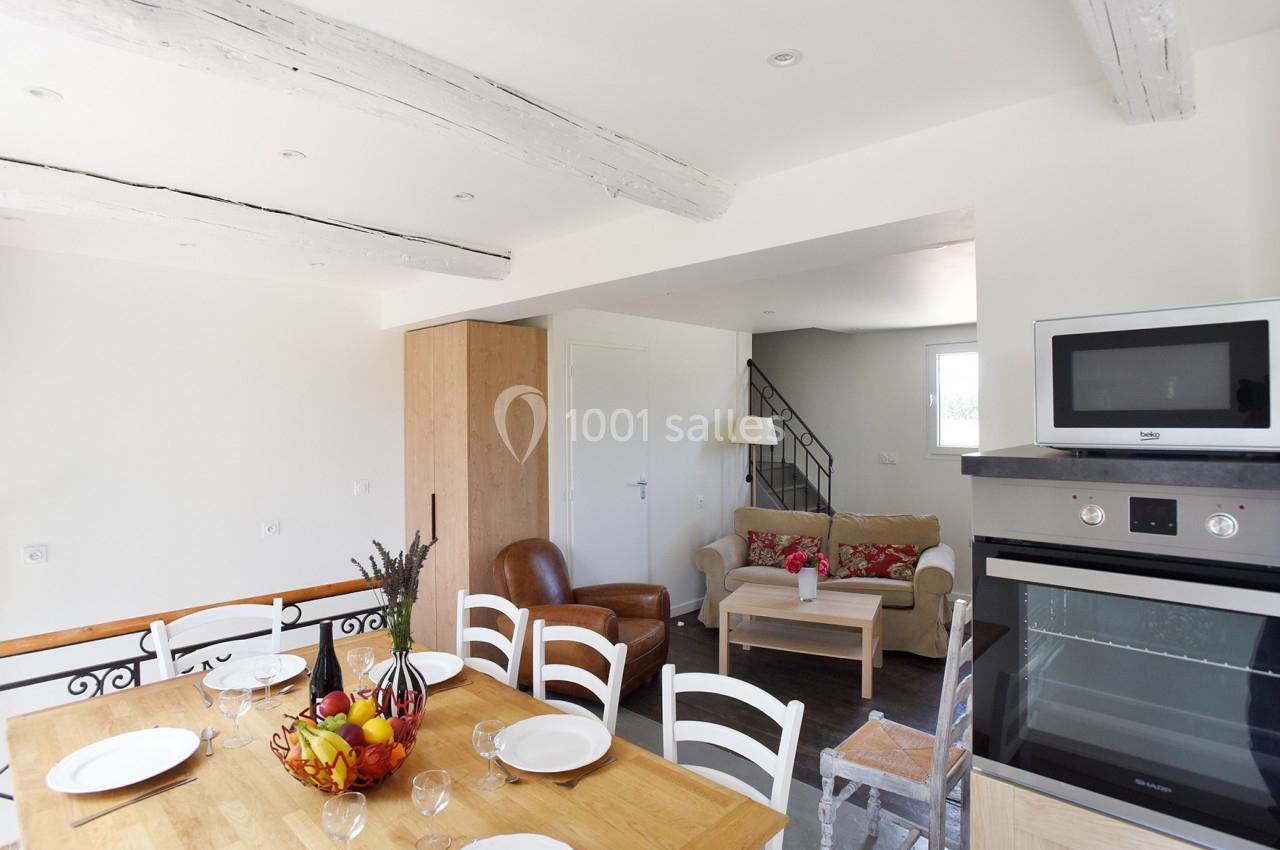 Salle à manger lumineuse avec table en bois, cuisine équipée et salon avec canapé, fauteuil et escalier en arrière-plan.
