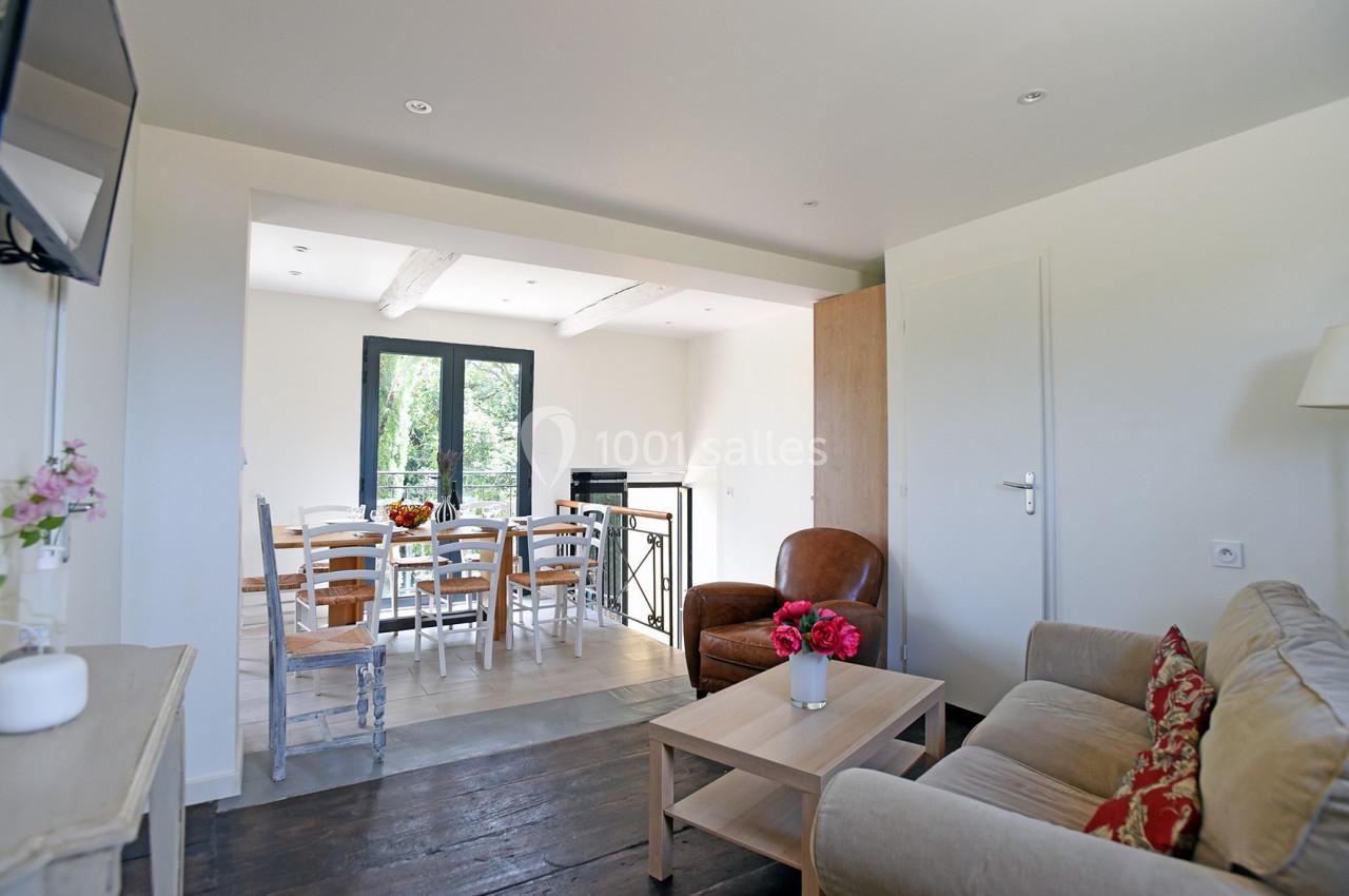 Salon lumineux avec canapé, fauteuil, table basse et vue sur une salle à manger avec table et chaises près d'une fenêtre.