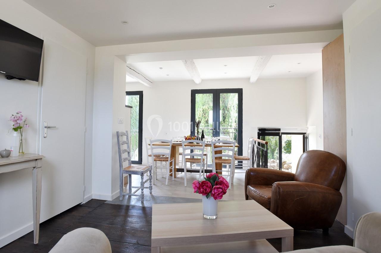 Salon lumineux avec canapé, fauteuil en cuir, table basse et vue sur une salle à manger avec table dressée.