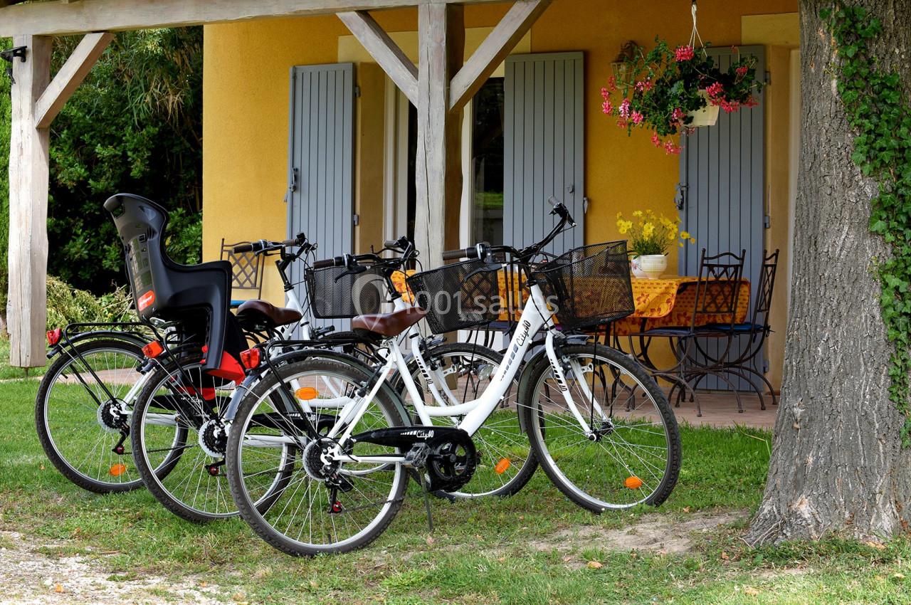 Trois vélos stationnés devant une maison avec des volets bleus, une terrasse et une table de jardin.